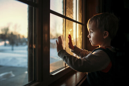 Boy looking through window while standing in darkroom at home - CAVF43473