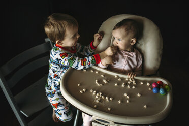 Bruder füttert seine Schwester mit Babynahrung, die auf einem Hochstuhl sitzt, in einer Dunkelkammer zu Hause - CAVF43479