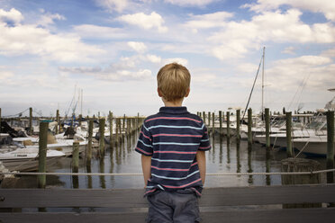 Rear view of boy standing on bridge by harbor against cloudy sky - CAVF43601