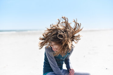 Happy girl shaking head while sitting at beach against sky - CAVF43641