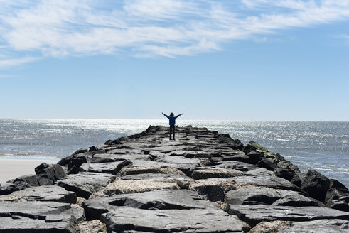 Rear view of girl with arms outstretched standing at rocky beach against sky during sunny day - CAVF43648