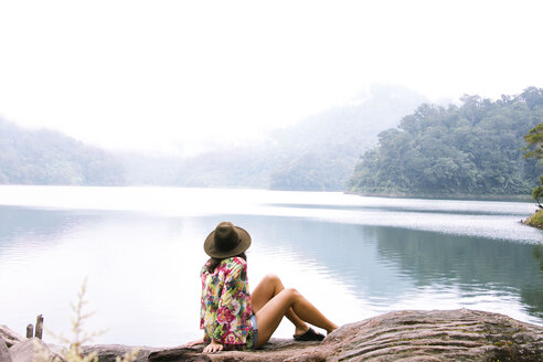 Side view of woman in hat sitting on log by lake during foggy weather - CAVF43674