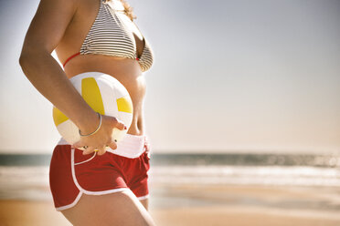 Midsection der jungen Frau hält Volleyball am Strand gegen klaren Himmel - CAVF43785