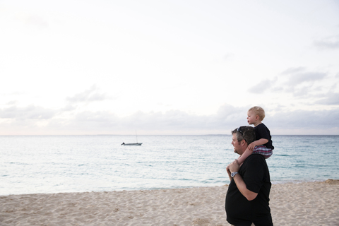 Glücklicher Vater, der seinen Sohn auf den Schultern trägt, während er am Strand steht, lizenzfreies Stockfoto