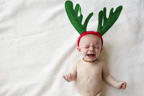 High angle view of cute baby boy in reindeer horns crying while lying on bed - CAVF43817