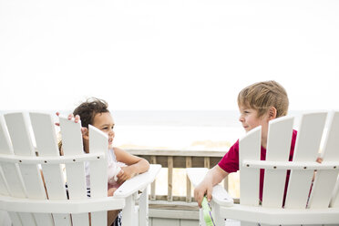 Glückliche Geschwister sitzen auf Liegestühlen am Strand gegen den klaren Himmel - CAVF43846