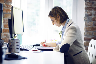 Businesswoman writing in book by computer at desk in office - CAVF43884