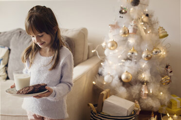 Glückliches Mädchen mit Plätzchen und Milchglas am Weihnachtsbaum zu Hause - CAVF43955