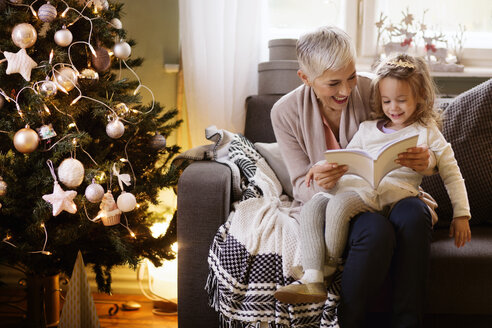 Großmutter und Mädchen lesen ein Buch, während sie zu Hause auf dem Sofa sitzen - CAVF43975