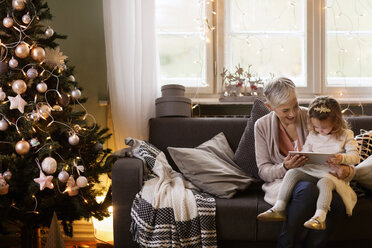 Großmutter hilft ihrer Enkelin bei der Benutzung eines Tablet-Computers, während sie auf dem Sofa sitzt - CAVF43984