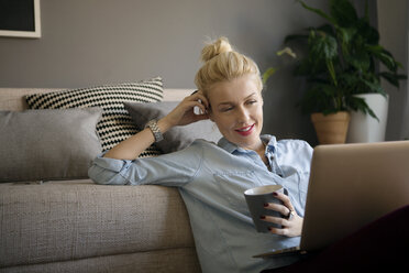 Lächelnde Frau, die eine Kaffeetasse hält und einen Laptop im Wohnzimmer benutzt - CAVF44059