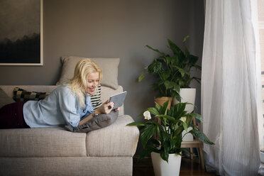 Glückliche Frau, die einen Tablet-Computer benutzt, während sie zu Hause auf dem Sofa liegt - CAVF44068