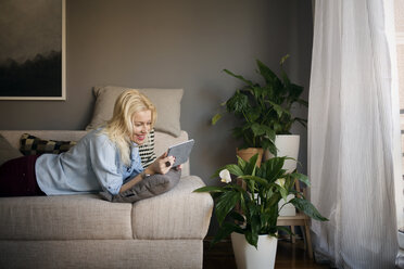 Happy woman using tablet computer while lying on sofa at home - CAVF44068