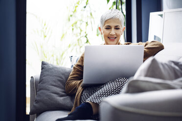 Smiling woman using laptop while sitting on sofa at home - CAVF44086