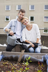 Portrait of young man holding watering can sitting with woman at urban garden - MASF06008