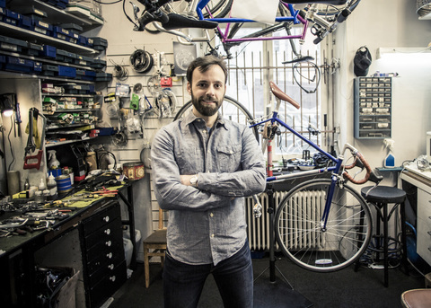 Porträt eines mittleren erwachsenen männlichen Besitzers, der mit verschränkten Armen in einer Fahrradwerkstatt steht, lizenzfreies Stockfoto