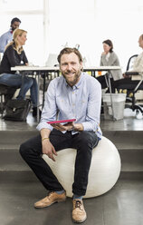 Portrait of mid adult businessman with digital tablet sitting on fitness ball in office - MASF06043