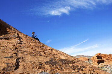 Niedriger Blickwinkel auf einen Jungen, der auf einem Felsen im Valley of Fire State Park vor blauem Himmel sitzt - CAVF44089