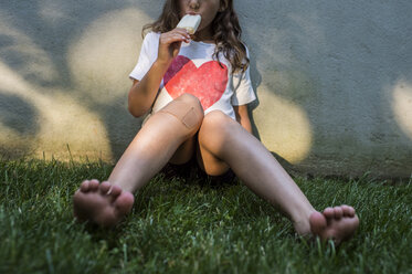 Girl eating ice cream while sitting on grassy field against wall in yard - CAVF44157