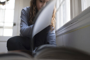 Mädchen liest ein Buch, während sie zu Hause am Fenster sitzt - CAVF44177