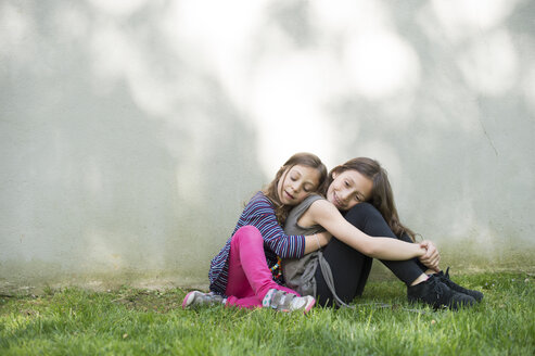 Seitenansicht eines Mädchens, das seine Schwester umarmt, während es im Hof an der Wand sitzt - CAVF44208
