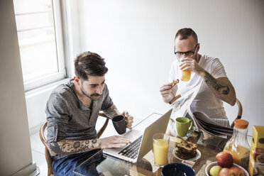 Schwule Männer, die einen Laptop benutzen und Zeitung lesen, während sie zu Hause frühstücken - CAVF44279