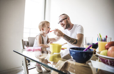 Glücklicher Vater füttert Tochter am Frühstückstisch vor hell erleuchteter Wand - CAVF44288