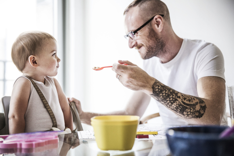 Glücklicher Vater füttert Tochter am Frühstückstisch an der Wand, lizenzfreies Stockfoto