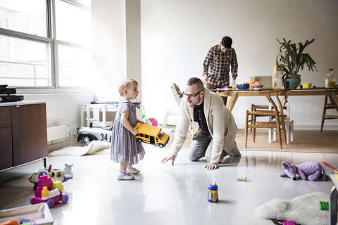 Vater spielt mit Tochter und Partnerin arbeitet am Tisch im hell erleuchteten Wohnzimmer, lizenzfreies Stockfoto