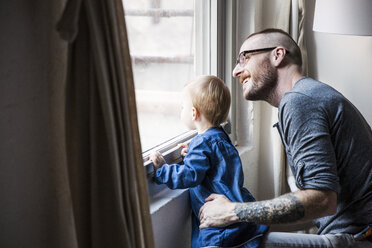 Glücklicher Vater mit Tochter, der durch das Fenster schaut, während er zu Hause sitzt - CAVF44308