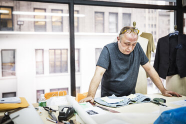 Serious fashion designer looking at jeans on table against glass window in design studio - CAVF44343