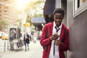 Lächelnder Student, der sein Telefon benutzt und sich an eine Wand in der Stadt lehnt - CAVF44351