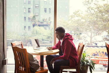 Student telefoniert beim Lernen am Glasfenster eines Restaurants - CAVF44354