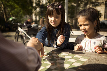 Schwestern spielen mit ihrem Großvater Dame, während sie an einem Tisch im Park sitzen - CAVF44356