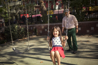 Grandfather looking at granddaughter enjoying on swing in park - CAVF44381