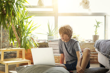 Junge schaut auf Laptop-Computer, während er auf dem Boden vor dem Fenster sitzt - CAVF44398