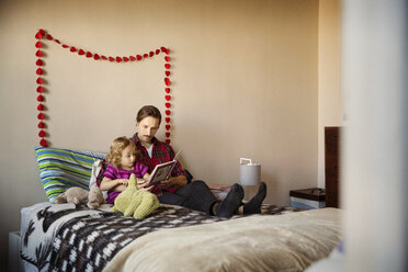 Vater zeigt einem Mädchen ein Buch, während es zu Hause auf dem Bett sitzt - CAVF44418