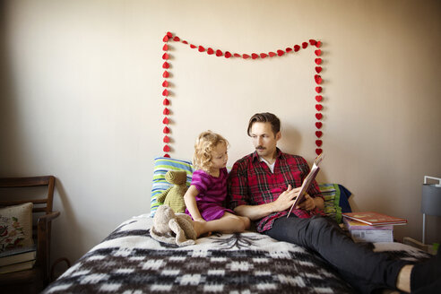 Vater zeigt seiner Tochter ein Buch, während sie zu Hause auf dem Bett an der Wand sitzt - CAVF44419