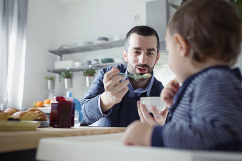 Vater füttert Sohn im Hochstuhl sitzend zu Hause mit Frühstück, lizenzfreies Stockfoto