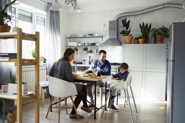 Parents looking at son sitting on high chair while having breakfast in kitchen - CAVF44484