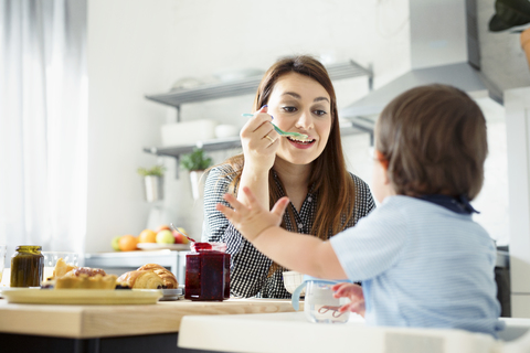 Mutter gibt ihrem Sohn am Küchentisch Frühstück, lizenzfreies Stockfoto