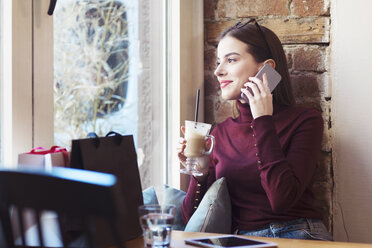 Frau mit Getränk, die durch das Fenster schaut, während sie in einem Café mit dem Handy telefoniert - CAVF44490