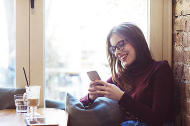 Glückliche Frau, die ein Mobiltelefon benutzt, während sie am Fenster eines Cafés sitzt - CAVF44496