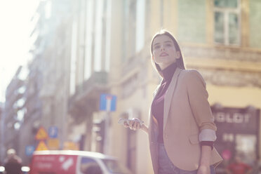 Woman holding mobile phone looking away while standing on city street - CAVF44501