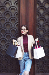 Happy woman with shopping bags looking away while standing by closed door - CAVF44517