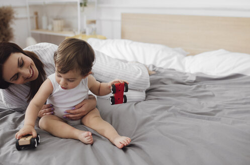 Happy mother looking at baby boy playing with toy cars on bed at home - CAVF44523