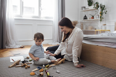 Glückliche Mutter schaut auf einen kleinen Jungen, der mit Spielzeug auf einem Teppich im Schlafzimmer spielt - CAVF44539