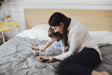Glückliche Mutter, die ihrem kleinen Jungen ein Bilderbuch zeigt, während sie zu Hause auf dem Bett sitzt - CAVF44541