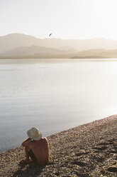 High angle view of shirtless man sitting at lakeshore against mountains and clear sky during sunny day - CAVF44572