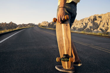 Low section of man holding skateboard while standing on country road on desert - CAVF44583
